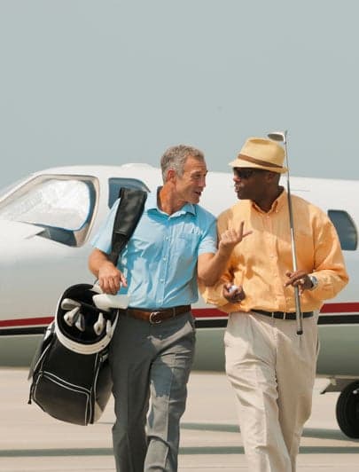 Two travelers carrying golf bags beside a private aircraft